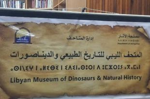 لوحة تعريفية تحمل اسم المتحف الليبي للتاريخ الطبيعي والديناصورات باللغتين العربية والإنجليزية، تتوسطها كتابة برموز قديمة، وتظهر عليها شعارات مصلحة الآثار وإدارة المتاحف، خلال فعالية أو عرض تعريفي داخل قاعة.