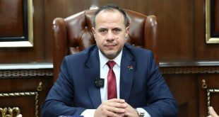 رجل يرتدي بدلة زرقاء وربطة عنق حمراء يجلس خلف مكتب على كرسي جلدي داخل مكتب رسمي، ويضع شارة علم ليبيا على سترته.