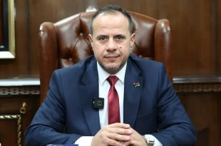 رجل يرتدي بدلة زرقاء وربطة عنق حمراء يجلس خلف مكتب على كرسي جلدي داخل مكتب رسمي، ويضع شارة علم ليبيا على سترته.