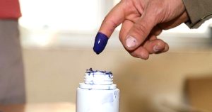 لقطة مقربة تُظهر إصبع سبابة لشخص يرتفع بعد غمسه في الحبر الانتخابي الفسفوري ذو اللون الأزرق الداكن، وتظهر أسفله فوهة زجاجة الحبر البيضاء البلاستيكية الملطخة ببقايا الحبر، مع خلفية ضبابية غير واضحة لمركز الاقتراع