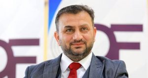 "لقطة متوسطة تظهر رجلاً ذا لحية خفيفة وشعر داكن قصير، يرتدي حلة رسمية رمادية (سترة وفيست) مع قميص أبيض وربطة عنق حمراء، ويبتسم بشكل خفيف أمام خلفية بيضاء تظهر بها أحرف باللون البنفسجي