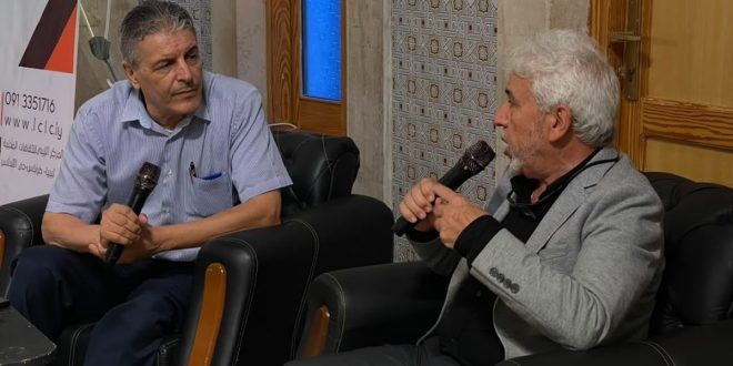 رجلان يتحدثان في ميكروفونات أثناء حوارية صحفية داخل بيت اسكندر