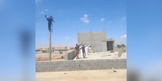 فني كهرباء يتسلق عموداً لإصلاح الأسلاك المقطوعة بمساعدة زملائه في منطقة سكنية قيد الإنشاء.