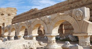 صف من الأعمدة والأقواس الرومانية الأثرية في الميدان السيفيري بمدينة لبدة الكبرى في ليبيا، تظهر فوقها منحوتات بارزة لرؤوس أسطورية (ميدوسا والنيريدات) داخل ميداليات رخامية تحت سماء صافية.