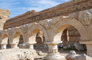 صف من الأعمدة والأقواس الرومانية الأثرية في الميدان السيفيري بمدينة لبدة الكبرى في ليبيا، تظهر فوقها منحوتات بارزة لرؤوس أسطورية (ميدوسا والنيريدات) داخل ميداليات رخامية تحت سماء صافية.