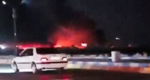 لقطة ليلية تظهر سيارة بيضاء متوقفة في مقدمة الصورة، وخلفها تصاعد لألسنة اللهب والدخان الأحمر الناجم عن قصف جوي استهدف مواقع في إيران.