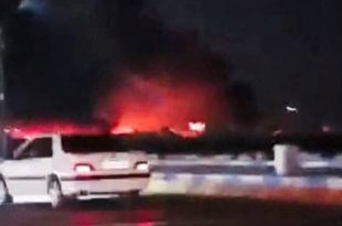 لقطة ليلية تظهر سيارة بيضاء متوقفة في مقدمة الصورة، وخلفها تصاعد لألسنة اللهب والدخان الأحمر الناجم عن قصف جوي استهدف مواقع في إيران.