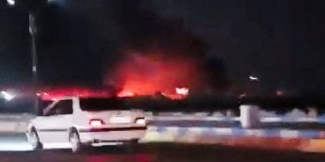 لقطة ليلية تظهر سيارة بيضاء متوقفة في مقدمة الصورة، وخلفها تصاعد لألسنة اللهب والدخان الأحمر الناجم عن قصف جوي استهدف مواقع في إيران.
