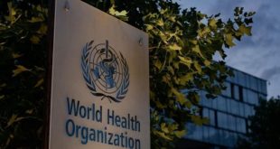 ​لوحة تحمل شعار واسم منظمة الصحة العالمية (World Health Organization) باللغتين الإنجليزية والعربية أمام مبنى المنظمة، وتحيط بها أشجار خضراء تحت سماء غائمة جزئياً