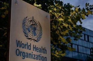 ​لوحة تحمل شعار واسم منظمة الصحة العالمية (World Health Organization) باللغتين الإنجليزية والعربية أمام مبنى المنظمة، وتحيط بها أشجار خضراء تحت سماء غائمة جزئياً