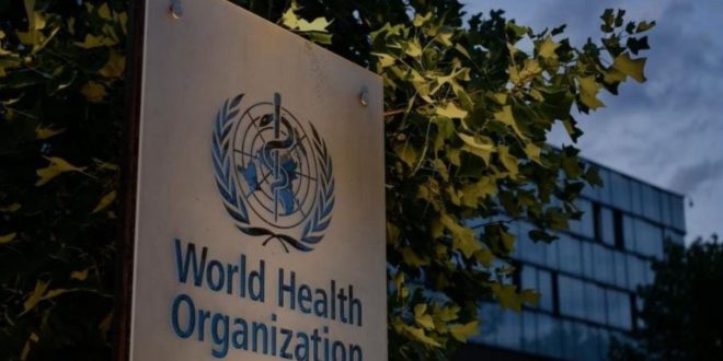 ​لوحة تحمل شعار واسم منظمة الصحة العالمية (World Health Organization) باللغتين الإنجليزية والعربية أمام مبنى المنظمة، وتحيط بها أشجار خضراء تحت سماء غائمة جزئياً