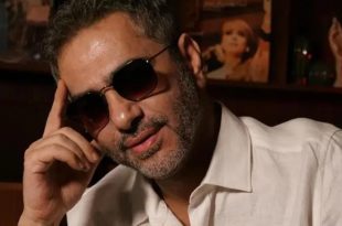 ​صورة مقربة للفنان اللبناني فضل شاكر يرتدي قميصاً أبيض ونظارة شمسية داكنة، ويضع يده على رأسه في لقطة تعبيرية، وخلفه جدار خشبي تظهر فيه ملامح لوحة أو صورة فنية