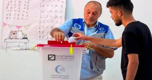 : شاب ليبي يقوم بإدخال ورقة الاقتراع داخل صندوق انتخابات بلاستيكي أبيض ذو غطاء أحمر، يحمل شعار "المفوضية الوطنية العليا للانتخابات" و"اللجنة المركزية لانتخابات المجالس المحلية للشباب"، ويقف بجانبه موظف مركز الاقتراع يرتدي سترة زرقاء لمساعدته، وتظهر في الخلفية سبورة مدرسية عليها جداول ضرب حسابية