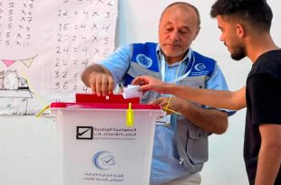 : شاب ليبي يقوم بإدخال ورقة الاقتراع داخل صندوق انتخابات بلاستيكي أبيض ذو غطاء أحمر، يحمل شعار "المفوضية الوطنية العليا للانتخابات" و"اللجنة المركزية لانتخابات المجالس المحلية للشباب"، ويقف بجانبه موظف مركز الاقتراع يرتدي سترة زرقاء لمساعدته، وتظهر في الخلفية سبورة مدرسية عليها جداول ضرب حسابية