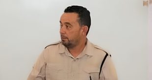 : لقطة متوسطة لرئيس قسم السلامة الوطنية في مدينة جادو، السيد خالد ساسي، يرتدي زي العمل الرسمي الفاتح (البيج) ويظهر بنظرة جانبية مستمعاً أو متحدثاً، على خلفية بيضاء سادة