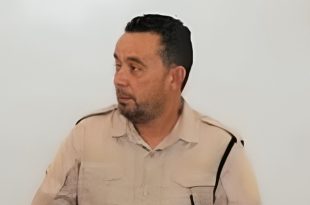 : لقطة متوسطة لرئيس قسم السلامة الوطنية في مدينة جادو، السيد خالد ساسي، يرتدي زي العمل الرسمي الفاتح (البيج) ويظهر بنظرة جانبية مستمعاً أو متحدثاً، على خلفية بيضاء سادة