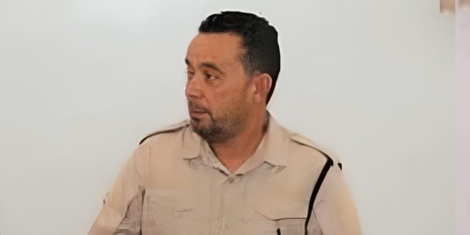 : لقطة متوسطة لرئيس قسم السلامة الوطنية في مدينة جادو، السيد خالد ساسي، يرتدي زي العمل الرسمي الفاتح (البيج) ويظهر بنظرة جانبية مستمعاً أو متحدثاً، على خلفية بيضاء سادة