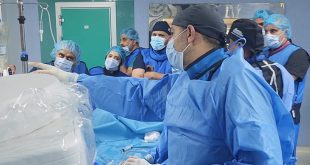 ​لقطة داخل غرفة العمليات بالمستشفى الجامعي طرابلس تُظهر فريقاً طبياً يرتدي الملابس الزرقاء والكمامات والملابس الواقية، حيث يقف طبيب في المقدمة يشير بيده نحو الأجهزة الطبية المغطاة ببلاستيك معقم، بينما يتابعه بقية الأطباء والممرضين باهتمام أثناء إجراء عملية زراعة الصمام الأورطي بالقسطرة