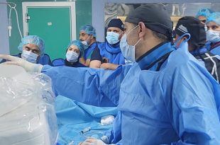​لقطة داخل غرفة العمليات بالمستشفى الجامعي طرابلس تُظهر فريقاً طبياً يرتدي الملابس الزرقاء والكمامات والملابس الواقية، حيث يقف طبيب في المقدمة يشير بيده نحو الأجهزة الطبية المغطاة ببلاستيك معقم، بينما يتابعه بقية الأطباء والممرضين باهتمام أثناء إجراء عملية زراعة الصمام الأورطي بالقسطرة