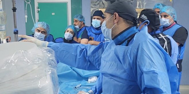 ​لقطة داخل غرفة العمليات بالمستشفى الجامعي طرابلس تُظهر فريقاً طبياً يرتدي الملابس الزرقاء والكمامات والملابس الواقية، حيث يقف طبيب في المقدمة يشير بيده نحو الأجهزة الطبية المغطاة ببلاستيك معقم، بينما يتابعه بقية الأطباء والممرضين باهتمام أثناء إجراء عملية زراعة الصمام الأورطي بالقسطرة