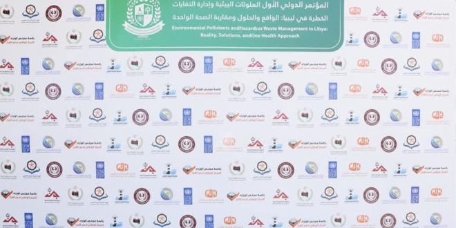​لوحة خلفية بيضاء كبيرة خاصة بالملتقى البيئي الأول في طرابلس، يظهر في أعلاها لافتة خضراء مكتوب عليها باللغتين العربية والإنجليزية "المؤتمر الدولي الأول لإدارة الملوثات البيئية والنفايات الخطرة في ليبيا"، وتتكرر على بقية اللوحة شعارات الجهات المشاركة والمنظمة بشكل منتظم.