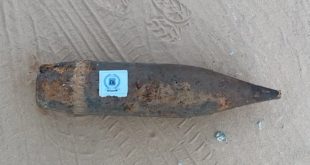 ​لقطة من الأعلى لقذيفة مدفعية كبيرة الحجم، قديمة ويظهر عليها الصدأ، مستلقية على أرض رملية تظهر عليها آثار إطارات، ويوجد ملصق صغير مربع يحمل شعاراً أمنياً (جهاز المباحث الجنائية الليبي) في منتصف القذيفة.
