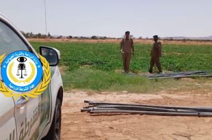 ​جانب من سيارة دورية تابعة لجهاز الشرطة الزراعية الليبية يظهر عليها شعار وزارة الزراعة والثروة الحيوانية، وفي الخلفية يظهر عنصران من أفراد الأمن يرتديان الزي العسكري أثناء قيامهما بجولة تفتيشية داخل حقل زراعي أخضر في منطقة قصر بن غشير