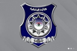 ​"شعار رسمي لوزارة الداخلية والشرطة الليبية يأتي على شكل درع باللون الأزرق الداكن والإطار الفضي، مكتوب في أعلاه 'وزارة الداخلية' وفي أسفله 'الشرطة'، ويتوسطه رمز نجمة وإكليل وشعار العين بألوان العلم الليبي، بخلفية رمادية."