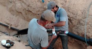 ​لقطة قريبة لعاملين من الشركة العامة للكهرباء يرتديان قبعات وملابس عمل، وهما يقومان بإصلاح وتوصيل أسلاك كابل كهربائي رئيسي سميك باللون الأسود داخل خندق ترابي محفور في الأرض أثناء أعمال الصيانة الطارئة