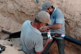 ​لقطة قريبة لعاملين من الشركة العامة للكهرباء يرتديان قبعات وملابس عمل، وهما يقومان بإصلاح وتوصيل أسلاك كابل كهربائي رئيسي سميك باللون الأسود داخل خندق ترابي محفور في الأرض أثناء أعمال الصيانة الطارئة