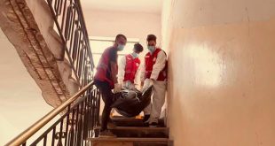 أعضاء من فريق إدارة الجثث بالهلال الأحمر الليبي يرتدون أزياء الحماية وستر حمارء ينزلون درج مبنى سكني وهم يحملون كيس جثامين.