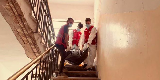 أعضاء من فريق إدارة الجثث بالهلال الأحمر الليبي يرتدون أزياء الحماية وستر حمارء ينزلون درج مبنى سكني وهم يحملون كيس جثامين.