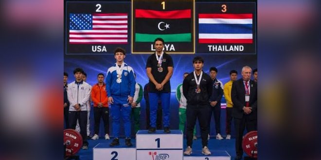 البطل الليبي محمد الساحلي يقف على منصة التتويج بالمركز الأول رافعًا علم ليبيا، وبجانبه الفائزان بالمركزين الثاني والثالث من الولايات المتحدة وتايلاند.