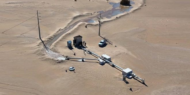لقطة جوية لمنشأة بئر مياه وأنابيب تدفق في وسط الصحراء مع وجود أعمدة كهرباء ومركبة خدمة بيضاء بجوارها.