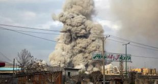 تصاعد عمود كثيف جداً من الدخان والتراب الرمادي في السماء إثر انفجار ضخم خلف مبانٍ ولائحة إعلانية مكتوبة بالفارسية.