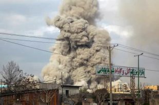 تصاعد عمود كثيف جداً من الدخان والتراب الرمادي في السماء إثر انفجار ضخم خلف مبانٍ ولائحة إعلانية مكتوبة بالفارسية.