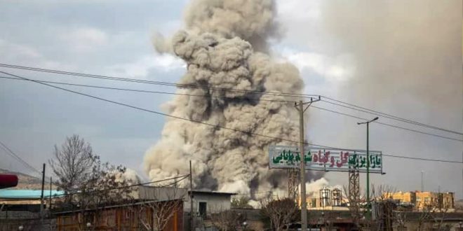 تصاعد عمود كثيف جداً من الدخان والتراب الرمادي في السماء إثر انفجار ضخم خلف مبانٍ ولائحة إعلانية مكتوبة بالفارسية.