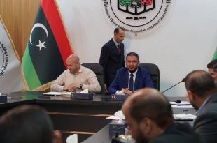 صورة لاجتماع رسمي داخل قاعة مؤتمرات، حيث يجلس وزير الزراعة الليبي ومسؤولون آخرون حول طاولة، خلفهم أعلام ليبيا وشعار المركز الوطني للوقاية والحجر الزراعي. يظهر المسؤولون بملابس رسمية ويشاركون في نقاش حول تطوير القطاع الزراعي.
