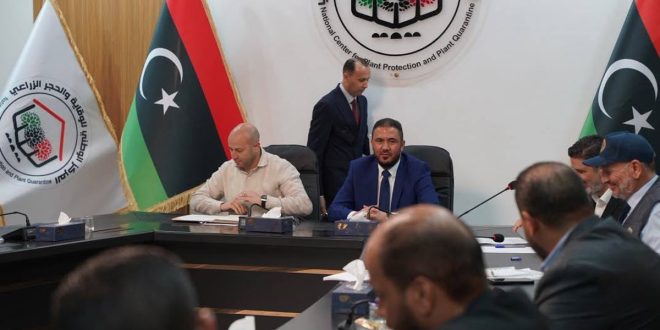 صورة لاجتماع رسمي داخل قاعة مؤتمرات، حيث يجلس وزير الزراعة الليبي ومسؤولون آخرون حول طاولة، خلفهم أعلام ليبيا وشعار المركز الوطني للوقاية والحجر الزراعي. يظهر المسؤولون بملابس رسمية ويشاركون في نقاش حول تطوير القطاع الزراعي.
