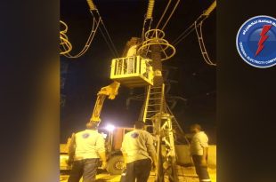 عنصر فني يتواجد داخل سلة رافعة هيدروليكية لإجراء أعمال صيانة على أبراج ومعدات الضغط العالي ليلاً بمحطة أوباري، ويقف في الأسفل زملاؤه يتابعون الأعمال، مع وجود شعار الشركة العامة للكهرباء.