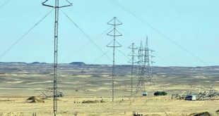 لقطة نهارية تظهر عدة أبراج وخطوط لنقل الطاقة الكهربائية ممتدة عبر منطقة صحراوية مفتوحة، مع وجود بعض الآليات وسيارات العمل الفنية في الموقع لاستكمال المشروعات المعتمدة للشركة العامة للكهرباء.