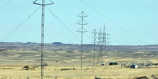 لقطة نهارية تظهر عدة أبراج وخطوط لنقل الطاقة الكهربائية ممتدة عبر منطقة صحراوية مفتوحة، مع وجود بعض الآليات وسيارات العمل الفنية في الموقع لاستكمال المشروعات المعتمدة للشركة العامة للكهرباء. خط قمرا (2)