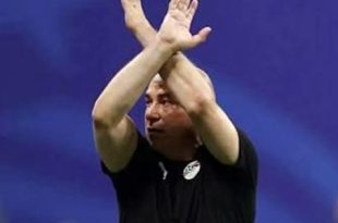 لقطة للمدير الفني لمنتخب مصر يرفع ذراعيه متقاطعتين على شكل حرف X (إشارة مكافحة العنصرية) أثناء مباراة، مرتدياً قميصاً أسود يحمل شعار المنتخب، والخلفية زرقاء.