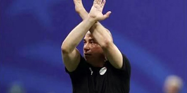 لقطة للمدير الفني لمنتخب مصر يرفع ذراعيه متقاطعتين على شكل حرف X (إشارة مكافحة العنصرية) أثناء مباراة، مرتدياً قميصاً أسود يحمل شعار المنتخب، والخلفية زرقاء.
