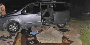 تُظهر الصورة مركبة فضية متوقفة في منطقة خارجية خلال الليل، وبابها الجانبي مفتوح. في المقدمة يظهر شخص مستلقٍ على بطانية فوق الأرض ومغطى بغطاء، بينما يبدو شخص آخر جالسًا داخل المركبة. تنتشر بعض الأغراض الشخصية والملابس حول مكان الاستراحة، ويعكس المشهد توقفًا أو مبيتًا مؤقتًا في موقع مفتوح.