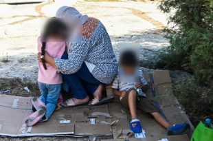 امرأة تحتضن طفلًا وتقف بجوار طفل آخر يجلس على قطعة كرتون في منطقة خارجية، مع تمويه وجوه الأشخاص للحفاظ على الخصوصية.