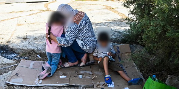 امرأة تحتضن طفلًا وتقف بجوار طفل آخر يجلس على قطعة كرتون في منطقة خارجية، مع تمويه وجوه الأشخاص للحفاظ على الخصوصية.