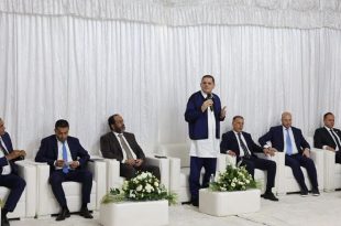 ​رئيس حكومة الوحدة الوطنية عبد الحميد الدبيبة يلقي كلمة واقفاً وممسكاً بمذياع، خلال اجتماعه مع أعيان وحكماء بلدية زليتن بحضور عدد من الوزراء والمسؤولين الجالسين على مقاعد بيضاء خلف خلفية من الستائر البيضاء