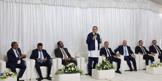 ​رئيس حكومة الوحدة الوطنية عبد الحميد الدبيبة يلقي كلمة واقفاً وممسكاً بمذياع، خلال اجتماعه مع أعيان وحكماء بلدية زليتن بحضور عدد من الوزراء والمسؤولين الجالسين على مقاعد بيضاء خلف خلفية من الستائر البيضاء