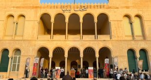 ​لقطة جماعية لافتتاح المعرض الوطني للفنون التشكيلية "رؤى معاصرة" أمام واجهة مبنى قصر الخلد الأثري في طرابلس، يظهر فيها الحضور والمنظمون على منصة أمام الأقواس التاريخية للمبنى مع وجود كاميرات تصوير وتغطية إعلامية وشعار منصة الصباح
